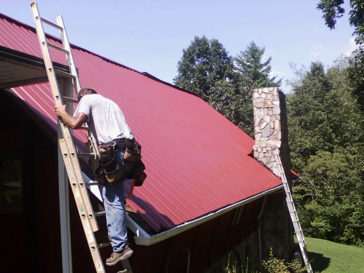 Roofer on a ladder beside a red metal roof and stone chimney — Soffit & Fascia Repair by Roofing Brothers in Dunn